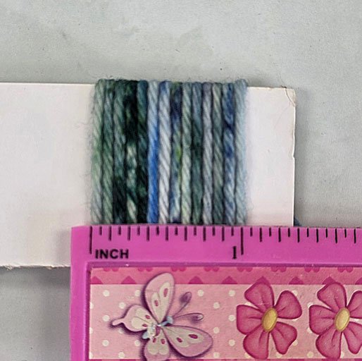 Demonstration of measuring WPI - yarn is wrapped around a piece of card with a ruler placed over the top showing 14 wraps measure one inch.