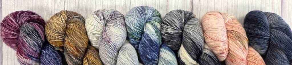 Row of 7 skeins of single ply yarns. All skeins are variegated hand dyed colourways